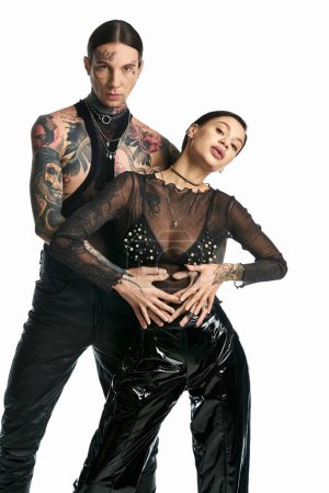 A young, tattooed man and woman in black outfits stand close together in a stylish pose against a grey studio background.