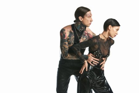 A young, tattooed couple in sleek black clothing poses in a studio setting against a grey background.