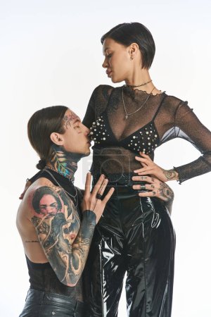 Un jeune couple élégant avec des tatouages debout en toute confiance dans un studio sur un fond gris.