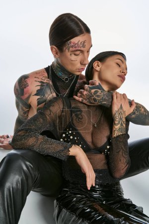A young, tattooed man and woman sit closely together in a studio, exuding style and connection on a grey background.