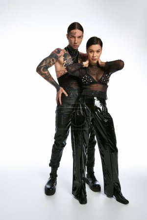 A stylish and tattooed couple stands together in a studio against a grey background, showcasing unity and affection.