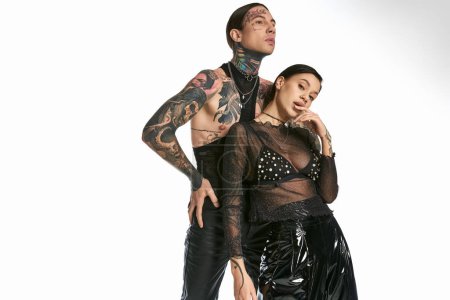 A stylish man with tattoos stands next to a woman in a captivating black dress in a studio against a grey background.
