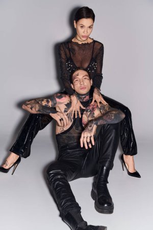 A stylish young man with tattoos sits on the back of a woman in a studio against a grey background.