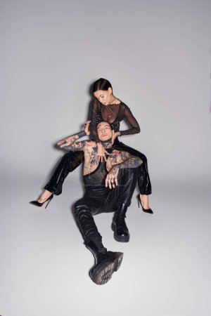 A young stylish woman is seated on top of a man in a studio, both covered in tattoos, against a grey background.