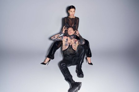 A stylish, tattooed couple in a studio setting, the woman sitting on top of the man against a grey background.