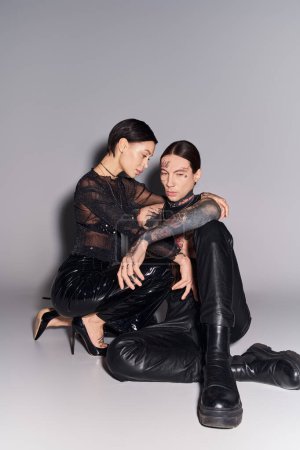 A young, stylish, and tattooed man and woman sitting closely together on a grey studio background.