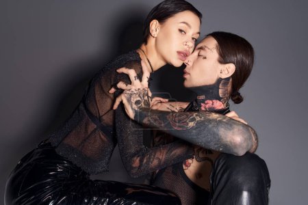 Two young stylish women with tattoos sitting closely together in a studio against a grey background.