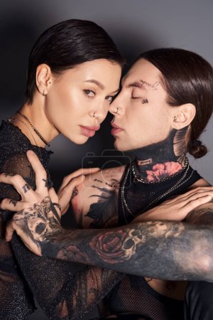 A stylish, tattooed couple sharing a warm hug in a studio against a grey background, expressing love and intimacy.