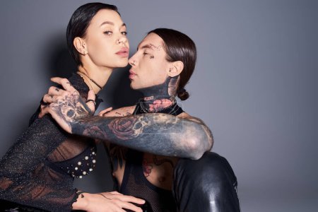 A young, stylish, tattooed man and woman strike a pose in a studio against a grey background.