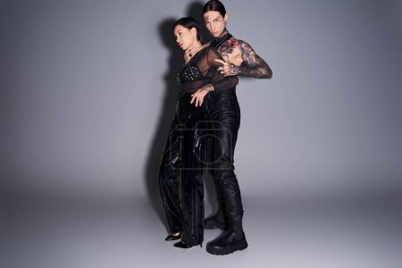 A young, stylish, and tattooed couple standing together in a studio against a grey background.
