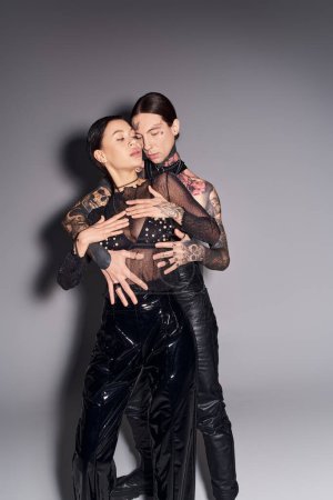 A young stylish couple with tattoos embrace each other warmly in a studio against a grey background.