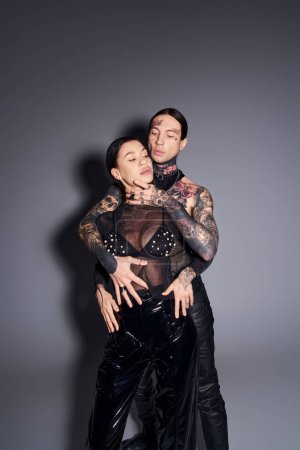 Una pareja joven y elegante con tatuajes vestidos con trajes de cuero negro a juego posan en un estudio sobre un fondo gris.