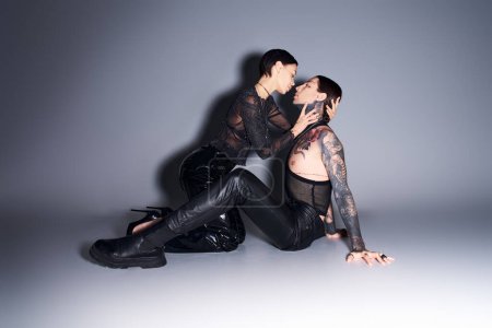 Foto de Una pareja joven, elegante y tatuada se relaja en el suelo en un estudio sobre un fondo gris. - Imagen libre de derechos