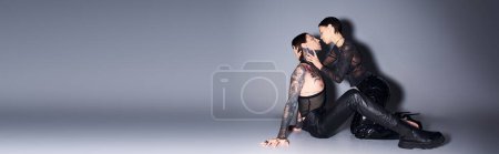 Foto de Una mujer joven, elegante y tatuada, se sienta con gracia en el suelo cerca del hombre en un estudio sobre un fondo gris. - Imagen libre de derechos