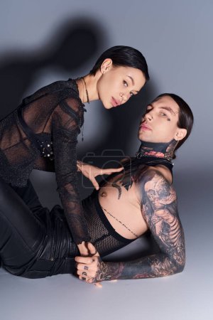 Un jeune homme et une jeune femme élégants avec des tatouages posant ensemble dans un studio sur un fond gris.