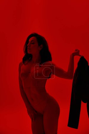 A captivating woman stands in a red room wearing lingerie, exuding allure.