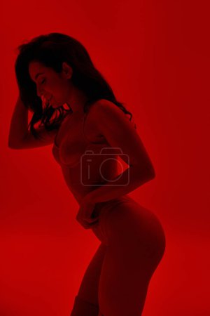 Téléchargez les photos : Femme en lingerie se tient avec confiance avec les mains sur les hanches dans une salle rouge vibrante. - en image libre de droit