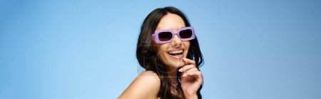 Téléchargez les photos : Une femme élégante dans une robe et des lunettes de soleil pose en toute confiance dans un studio avec fond bleu. - en image libre de droit