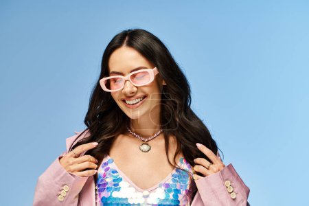 A fashionable woman radiates summertime vibes in a pink jacket and pink sunglasses against a blue studio backdrop.