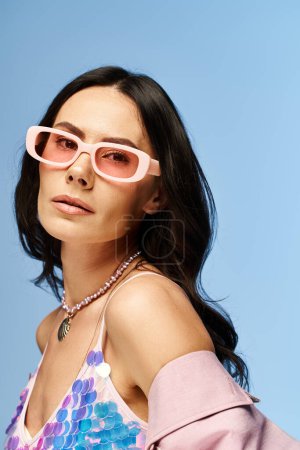A stylish woman wearing pink sunglasses and a pink top poses confidently against a blue studio background, exuding summer vibes.