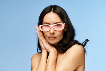 Foto de Una mujer con estilo está tomando una pose en gafas de sol rosadas contra un telón de fondo de estudio azul, que encarna vibraciones veraniegas. - Imagen libre de derechos