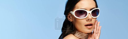 A pretty woman exudes charm wearing white sunglasses and a choker in a studio against a blue background, embodying summertime vibes.