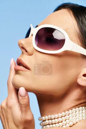 Téléchargez les photos : Une femme élégante avec des lunettes de soleil et des perles autour de son cou pose dans un studio sur un fond bleu vif. - en image libre de droit