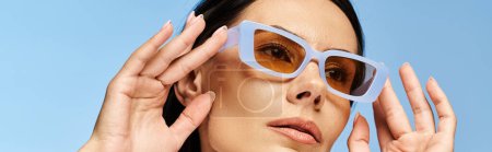 A stylish woman in sunglasses with hands on face, exuding coolness on a blue studio backdrop.