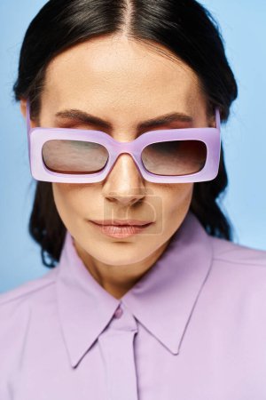 Foto de Una mujer de moda emana vibraciones veraniegas en una camisa púrpura y gafas de sol a juego contra un fondo de estudio azul. - Imagen libre de derechos