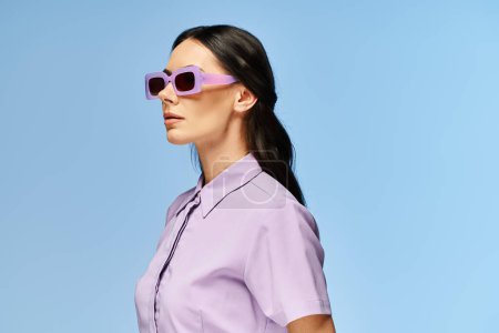 Eine stylische Frau mit Sonnenbrille und lila Hemd posiert selbstbewusst vor leuchtend blauem Studiohintergrund.