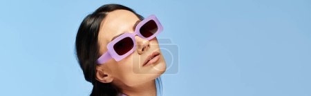 A stylish woman wearing sunglasses gazes up at the sky against a blue studio backdrop.