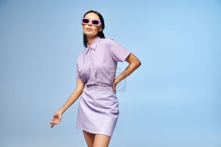 A beautiful woman in a purple dress and stylish sunglasses poses in a studio against a summery blue background.