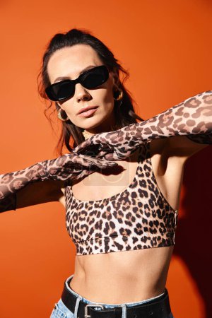 Eine stilvolle Frau strahlt Vertrauen in Leopardenmuster und Sonnenbrille vor leuchtend orangefarbenem Hintergrund aus.