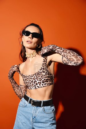 Eine modische Frau in Leopardenmuster und Sonnenbrille posiert selbstbewusst in einem Studio vor orangefarbenem Hintergrund.