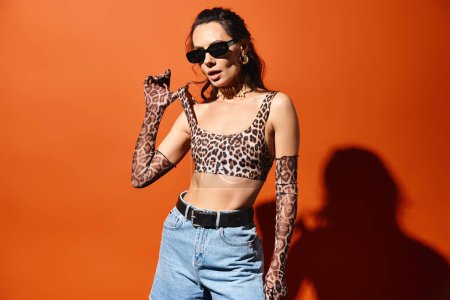Stylish woman in sunglasses posing confidently in a leopard print top and jeans against an orange background.