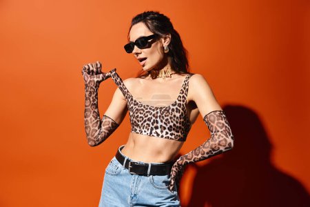 Eine modische Frau trägt selbstbewusst ein Oberteil mit Leopardenmuster und Jeans mit Sonnenbrille vor orangefarbener Studiokulisse.