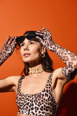 Eine stylische Frau im Leopardenmuster hält während eines sommerlichen Mode-Shootings in einem leuchtend orangefarbenen Atelier die Hände auf dem Kopf.