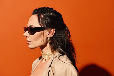 A stylish woman wearing sunglasses and a tan jacket poses against an orange background, exuding summertime fashion vibes.