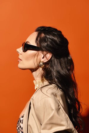 A stylish woman with sunglasses poses confidently in a fashionable jacket against an orange backdrop.