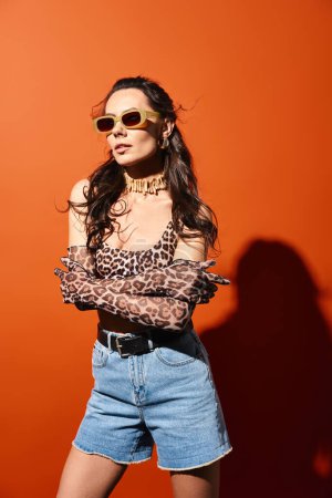 A stylish woman wearing leopard print top and denim shorts, exuding summertime vibes, on an orange background.