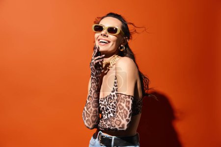 Foto de Una mujer con estilo mece un top de estampado de leopardo y gafas de sol, exudando encanto veraniego sobre un fondo naranja. - Imagen libre de derechos