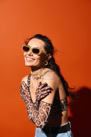 Eine stylische Frau präsentiert Sommermode in einem Leopardenmuster-Hemd und einer trendigen Sonnenbrille vor orangefarbenem Hintergrund.
