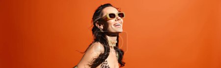 A stylish woman in a leopard print dress and sunglasses poses confidently in a studio against an orange background.
