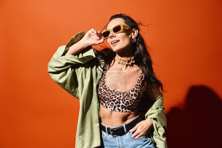 Une femme élégante portant un haut imprimé léopard et un jean, respirant la confiance et la mode estivale sur un fond de studio orange.