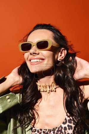 A stylish woman in a green jacket and sunglasses poses confidently against an orange background in a studio setting.