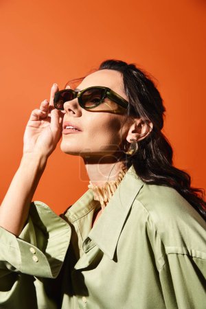 A stylish woman with sunglasses and a green shirt poses against an orange studio backdrop, exuding summertime fashion vibes.