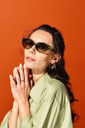A fashionable woman wearing sunglasses and a green shirt poses confidently in a studio with an orange background.