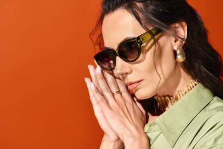 A pretty woman wearing stylish sunglasses, holding her hands to her face in a studio with an orange background.