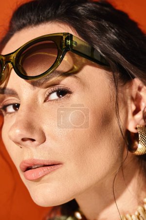 A stylish woman with sunglasses and a necklace poses in a studio with a vibrant orange background.