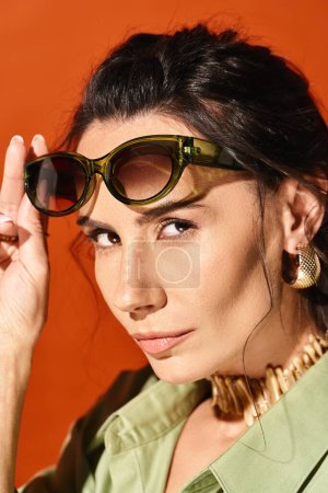 A fashionable woman adorned in a green shirt and sunglasses poses confidently in a studio with an orange background.
